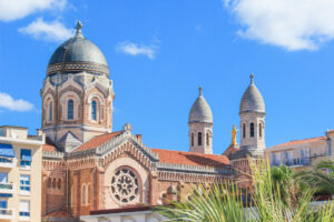 Basilika Notre-Dame de la Victoire in Saint-Raphaël mit Kuppel, Türmen und Palmen vor blauem Himmel
