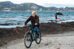 Radfahrer am Strand von Hyères mit Meer, Hügeln und Kitesurfern im Hintergrund