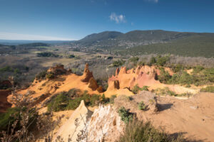 Blick über orangefarbene Ockerpfeiler auf das Tal und die bewaldeten Berge bei Rustrel